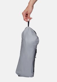 Main tenant une pochette grise à fermeture éclair par sa boucle en tissu noir sur un fond blanc uni.