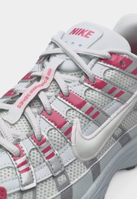 Baskets Nike blanches et grises avec des accents roses, détails en mesh, lacets superposés et logo "Nike" sur la languette et le côté.