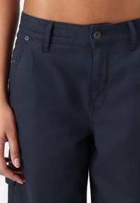 Marineblauwe broek met een rechte snit, voorzien van een knoopsluiting in de tailleband, voorzakken en een subtiele textuur. Gestikte details langs de naden.