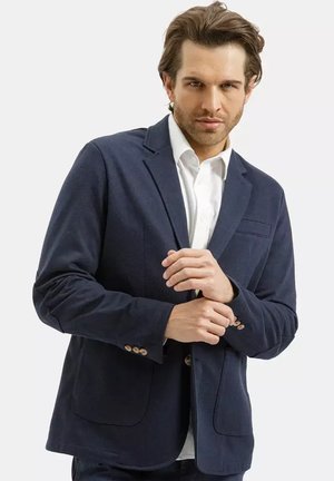 Mężczyzna poprawiający mankiety ciemnogranatowej marynarki noszonej na białej koszuli, zwrócony przodem, z neutralnym wyrazem twarzy na jednolitym tle.