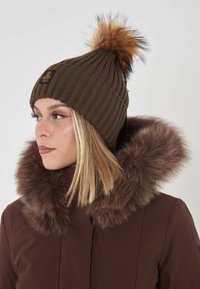Cappello a coste marrone con pompon in pelliccia sintetica sulla sommità, abbinato a un cappotto con cappuccio bordato di pelliccia marrone.