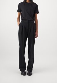 Person klädd i en svart t-shirt med korta ärmar och liten logotyp, högmidjade, plisserade svarta byxor och svarta skor, stående mot en enfärgad vit bakgrund.