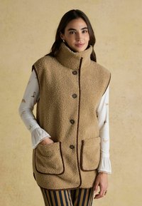 Beige Fleece-Weste mit hohem Kragen, braunen Akzenten und Knopfverschluss, ausgestattet mit zwei Fronttaschen. Langärmliges Shirt mit Tierdruck darunter.