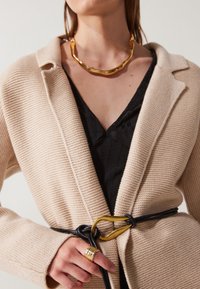 Beige gestrickter Blazer mit strukturiertem Finish, getragen über einem schwarzen Oberteil. Verfügt über einen schwarzen Gürtel mit goldenen Hardware-Akzenten und einem kräftigen goldenen Ring.
