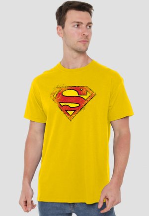 Man wearing yellow T-shirt with distressed red and yellow Superman logo, looking sideways against plain gray background.