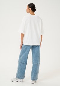 Camiseta blanca de gran tamaño con mangas cortas, combinada con jeans de pierna ancha de color azul claro. El modelo también lleva zapatillas blancas con detalles grises.