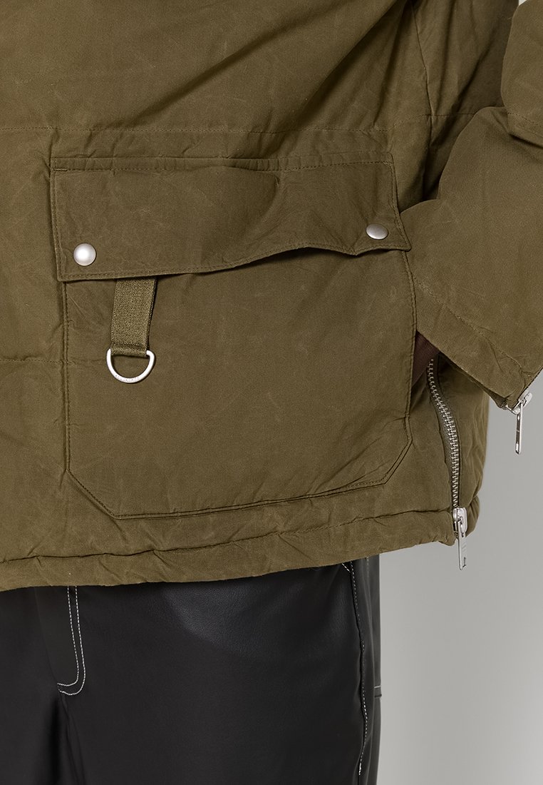 Zielona oliwkowa kurtka pikowana z zamkiem z boku, przednią kieszenią na klapie i metalowymi akcentami. Materiał wydaje się solidny, o miękkiej teksturze.