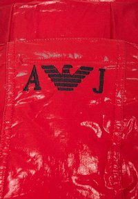 Röd tyg med en glänsande yta, med svarta broderade bokstäverna "A" och "J" och en stiliserad vinge-logotyp mellan dem.