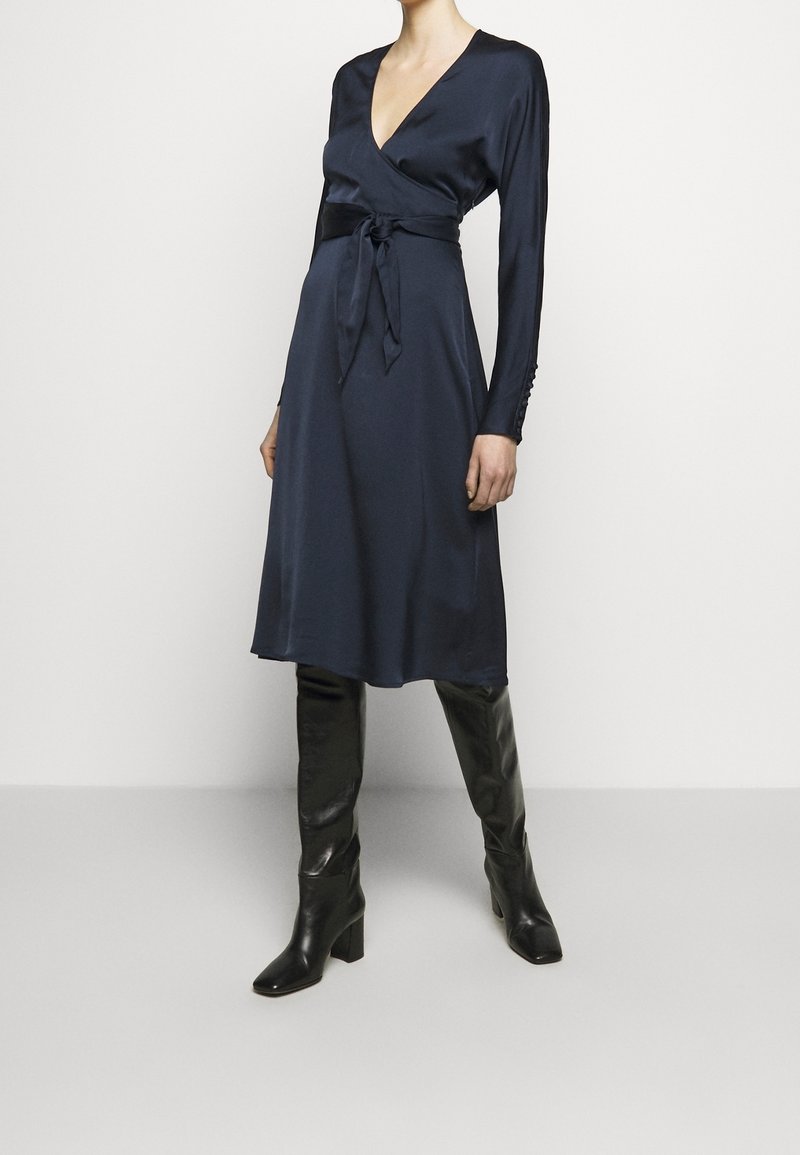 Robe portefeuille en satin bleu marine avec manches longues, décolleté plongeant en V, taille nouée et détails à boutons sur les poignets, associée à des bottes en cuir noir montantes jusqu'au genou.