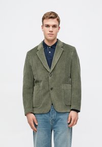 Maslinasto zelena kordura blazer s nazubljenim reverom, dva prednja džepa i dugmetom, nosi se preko tamnoplave polo majice i svijetlih traperica.