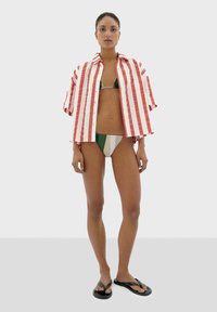 Rot- und weiß gestreifte Hemdbluse, die über einem grünen und cremefarbenen Bikini getragen wird. Das Model trägt schwarze Flip-Flops und steht vor einem neutralen Hintergrund.