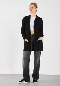 Zwarte geribde cardigan met lange mouwen, open voorkant en twee voorszakken, gecombineerd met hoog-waist grijze jeans en zwarte sneakers.