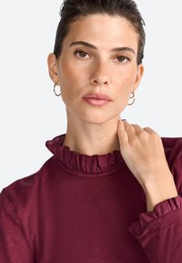 Burgundska bluza s ruffled izrezom i manžetama, izrađena od mekog materijala, s ležernim krojem i suptilnim sjajem.