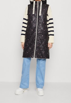 Czarna pikowana długa kamizelka z kapturem i zamkiem, noszona na paskowanym swetrze, jasnoniebieskich spodniach o szerokich nogawkach oraz białych butach na platformie.