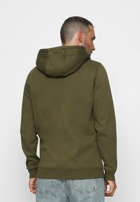 Hombre de espaldas vestido con una sudadera verde oliva con la capucha bajada y unos jeans de mezclilla azul claro contra un fondo liso.