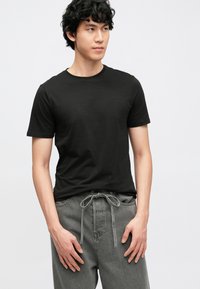 Črna bombažna majica s kratkimi rokavi in okroglim izrezom, kombinirana s sivimi hlačami z vrvico. Enostavna zasnova, gladka tekstura in sproščen kroj.