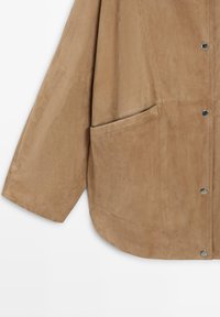 Béžová semišová bunda s dlouhými rukávy, velkou boční kapsou, patentními knoflíky a viditelnými švy.