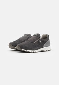 Pár tmavě šedých semišových sneakers bez šněrování se zipy na bocích a bílými gumovými podrážkami na čistém bílém pozadí.