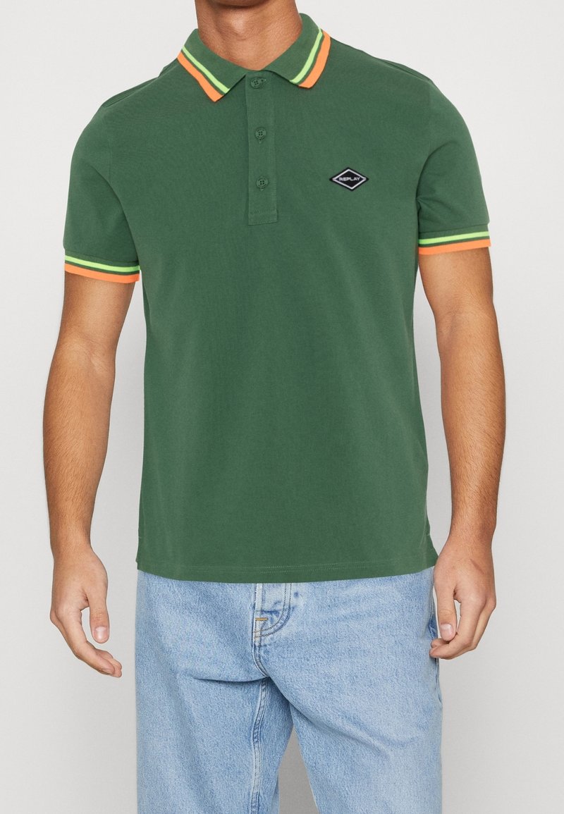 Homme portant un polo vert avec des rayures orange et jaune au col et aux manches, associé à un jean bleu clair.