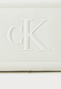 Borsa in pelle bianca con logo "CK" impresso, texture liscia e dettagli di cucitura sottili. Design compatto adatto a varie occasioni.
