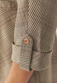 Tessuto a quadri marrone e beige con motivi variabili, caratterizzato da un bottone sul polsino e una texture morbida. Accenti blu intercalati.