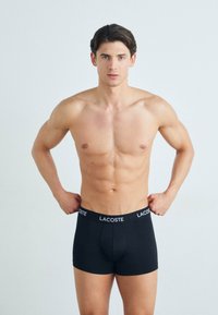 Boxer-briefs en coton noir avec un design ajusté et une ceinture élastique portant le logo "LACOSTE" en blanc. Texture simple et lisse.