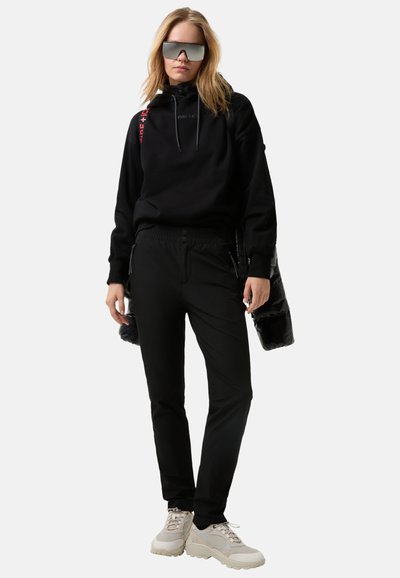 Schwarzer Hoodie mit Logo, schwarze, schmal geschnittene Hose und klobige beige Sneakers. Das Outfit wird mit einer verspiegelten Sonnenbrille und einer glänzenden schwarzen Jacke ergänzt.