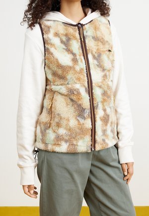 Osoba ubrana w polarową kamizelkę na zamek w wielokolorowych odcieniach beżu i brązu, na białej bluzie z kapturem, w jasnooliwkowych spodniach, stojąca na gładkim tle.