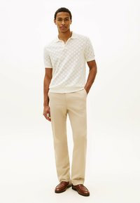 Giovane uomo in piedi con le mani in tasca, indossa una polo a maniche corte color crema con motivo, pantaloni beige e mocassini in pelle marrone su sfondo bianco.