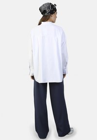 Camisa blanca de botonadura, de corte holgado, con cuello puntiagudo, combinada con pantalones anchos de color azul marino oscuro y un pañuelo negro con patrón de paisley blanco.