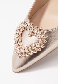 Metallic gold pump with a pointed toe, featuring a heart-shaped crystal embellishment on the vamp. Smooth leather texture.