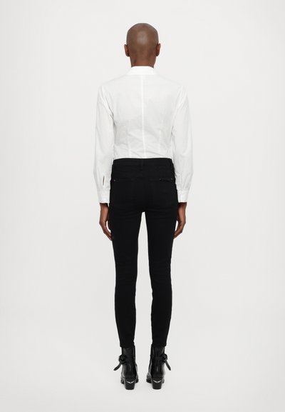 Camisa blanca de cuello con mangas largas, jeans skinny negros y botas de tobillo negras con acentos de lazo, vista desde la parte trasera. Texturas suaves.