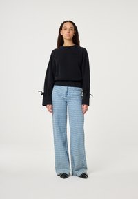 Zwarte cropped sweatshirt met wijde mouwen en strikaccenten, gecombineerd met lichtblauwe wijde jeans met een subtiel patroon. Zwarte schoenen.