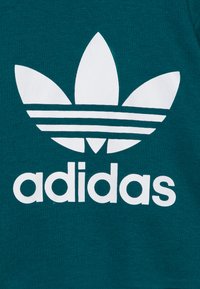 Logo trefoil bianco di Adidas e testo su sfondo in tessuto verde scuro.