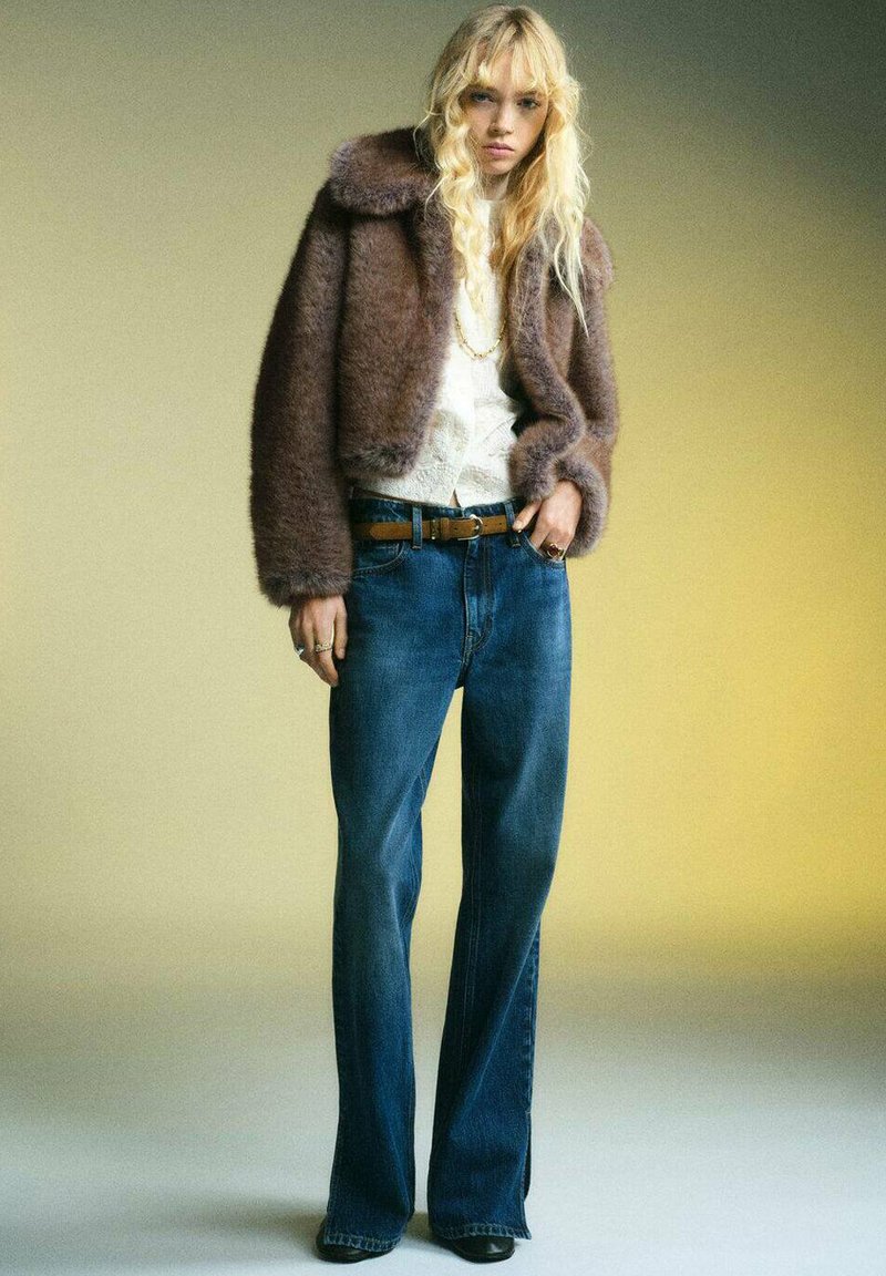Kroppad jacka i brun imitationspäls, vit texturerad blus, blå vida jeans, brunt läderbälte, svarta ankelstövlar, mot en gradientbakgrund.