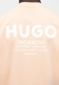 Άτομο που φοράει ανοιχτό μπεζ πουκάμισο με λευκό κείμενο τυπωμένο στο πάνω μέρος της πλάτης που γράφει «HUGO MCMXCIII Registered Trademark Since 1993».