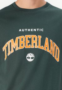 Camiseta de algodón verde oscuro con la palabra "AUTÉNTICO" en blanco y "TIMBERLAND" en negrita naranja arqueándose sobre un logo de árbol blanco.