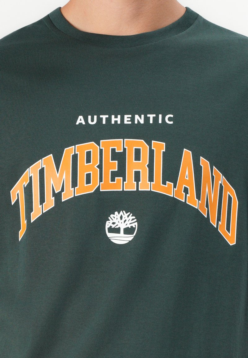 T-shirt in cotone verde scuro con la scritta "AUTHENTIC" in bianco e "TIMBERLAND" in arancione in grassetto che si cura sopra a un logo di albero bianco.