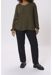 Femme portant un blouse à manches longues vert olive, un pantalon droit noir et des mocassins noirs avec une boucle dorée, se tenant sur un fond blanc.