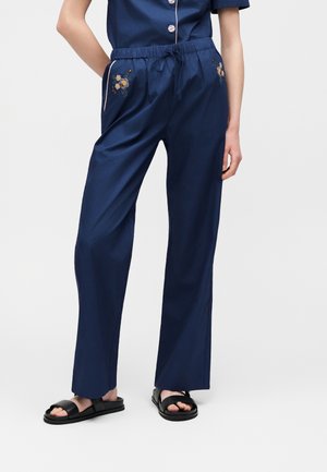 Asmuo dėvi tamsiai mėlynas pižamos kelnes su gėlėmis siuvinėtomis kišenėmis ir juodas šlepetes, stovintis su viena ranka kišenėje.