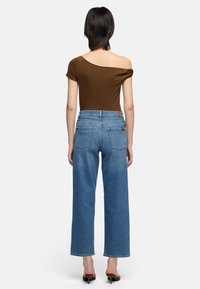 Femme debout de dos portant un haut marron à une épaule, un jean bleu à jambes droites et des sandales marron à talons sur fond blanc.