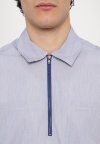 Camicia a righe di colore azzurro chiaro, con colletto corto e zip frontale con accenti blu. Include due tasche sul petto e una texture liscia.
