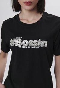 Camiseta de algodón negra con un slogan texturizado en lentejuelas plateadas "#BOSSIN" y el texto blanco "INFLUENCER Voy a lograrlo" debajo.