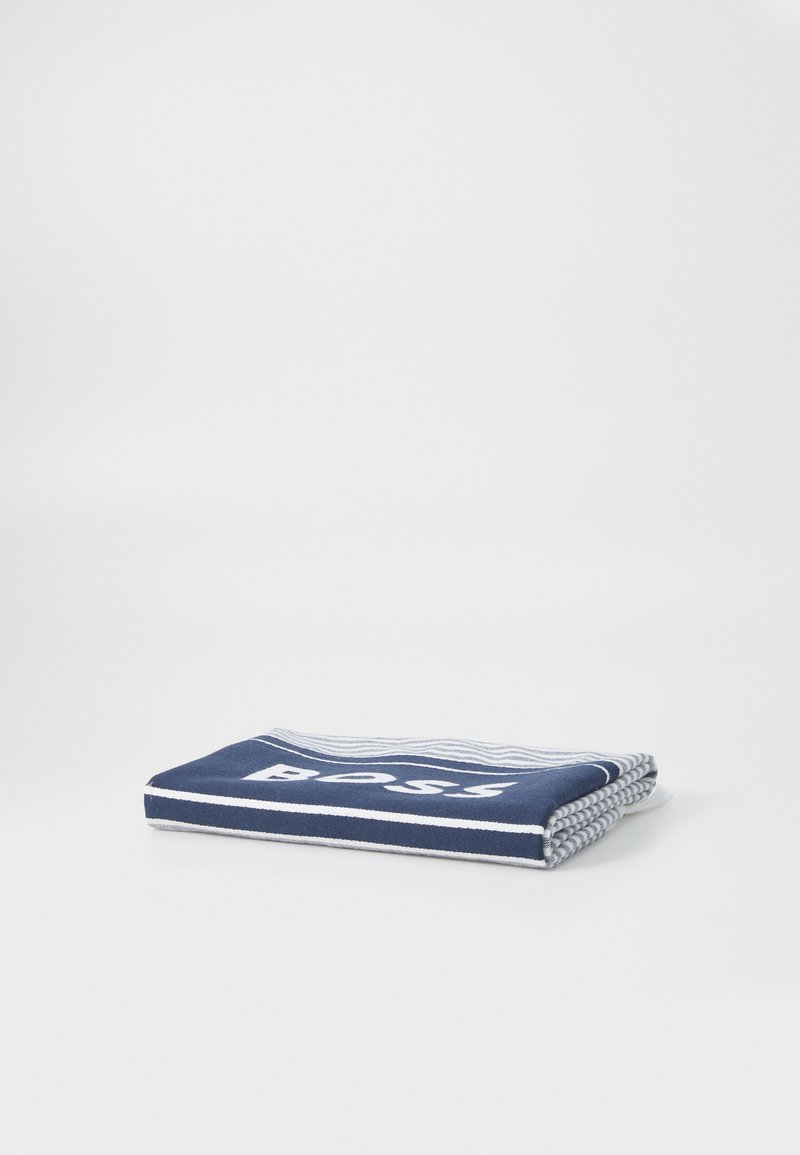 Σκούρο μπλε πετσέτα με λευκές ρίγες και λογότυπο, φτιαγμένη από μαλακό υλικό. Διπλωμένη προσεκτικά για να δείξει τη υφή και τις λεπτομέρειες του σχεδίου.