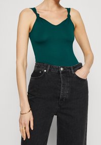 Teal ärmlös bodysuit med knutna axelremmar, i kombination med höga svarta jeans. Slätt tyg med en figurnära silhuett.