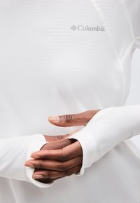 Χέρια με σκουρόχρωμο δέρμα που φορούν λευκό μακρυμάνικο πουκάμισο Columbia με τρύπες για τους αντίχειρες, εμφανίζοντας υφή υφάσματος και λογότυπο στο στήθος.