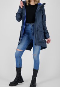 Wodoszczelna granatowa kurtka z niebieską prążkowaną podszewką, przednimi kieszeniami i kapturem na sznurku, połączona z niebieskimi podartymi dżinsami i czarnymi botkami.