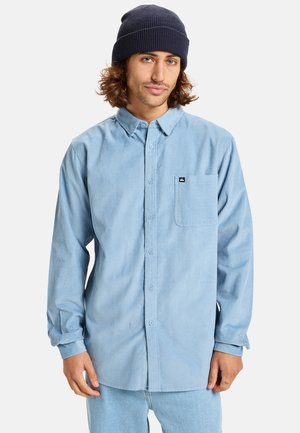 Light blue button-up shirt made of textured fabric, featuring a chest pocket and round collar, worn with a navy beanie.