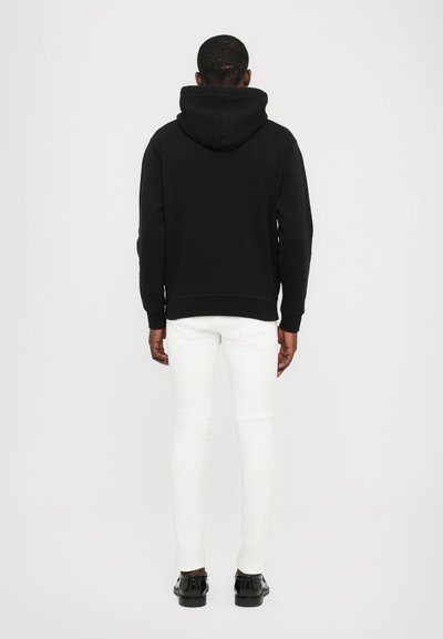 Felpa nera con cappuccio, polsini e orlo a coste; abbinata a jeans skinny bianchi e scarpe nere. Outfit semplice e informale.