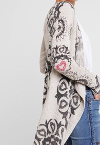 Beige open cardigan met zwarte bloemenpatronen, textuur gebreid stof en roze hartaccenten langs de mouwen en randen.